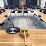 Clear image of a modern conference room with wooden tables and black chairs, equipped with microphones and presentation screens.